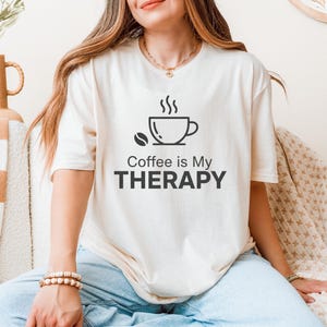 Op de afbeelding: Crèmekleurig T-shirt met de zwarte tekst "Coffee is My THERAPY". Het ontwerp bevat een koffiekopje met stoom en een koffieboon. Het T-shirt is een casual kledingstuk voor dagelijks gebruik.