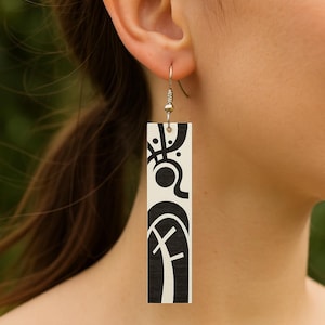 May include: A single earring with a rectangular shape and a white background. The earring features a black abstract design. The earring is hanging from a gold-colored hook. The earring is worn on the ear.