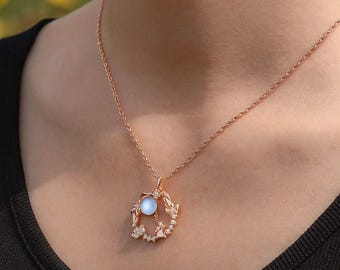 Peut inclure: Collier en or rose avec un pendentif orné d'un globe bleu clair et de délicats accents de feuilles et de cristaux. Le collier est présenté sur un fond sombre, mettant en valeur le design et la couleur des bijoux.