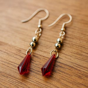 May include: A pair of gold-colored earrings with red, faceted, teardrop-shaped pendants. The earrings have a simple design with a gold hook, small gold beads, and a red crystal pendant. The earrings are displayed on a wooden surface.