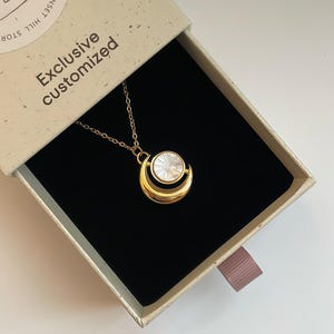 以下が含まれることがあります： 黒い内装の開いたギフトボックスに入った、円形ペンダントのゴールドネックレス。ペンダントは白く円形の中心部と金の三日月型をしています。ボックスはベージュで、「Exclusive customized」の文字が入っています。
