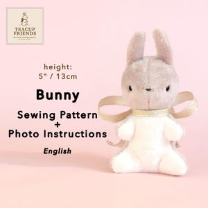 Puede incluir: Un patrón de costura de conejo de peluche con instrucciones fotográficas. El conejo es gris claro y blanco, con una cinta dorada alrededor del cuello. El conejo mide 13 cm de alto. El texto "Bunny Sewing Pattern + Photo Instructions English" es visible.