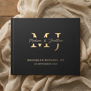 May include: A black hardcover book with gold lettering. The cover features the names "Madison & Jonathan" and the date "25 September 2025". The location "Brooklyn Botanic, NY" is also printed on the cover. The book is resting on a wooden surface with a beige cloth.