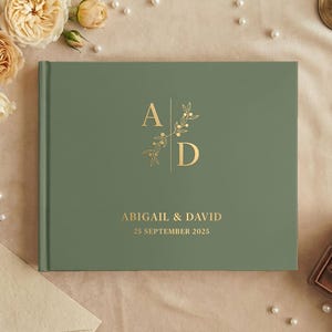 May include: A sage green hardcover guest book featuring gold foil lettering. The cover displays the initials A and D separated by a vertical line with a botanical sprig, followed by the names Abigail & David and the date 25 September 2025.