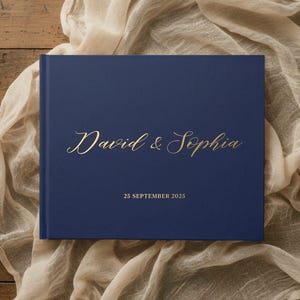 Puede incluir: Un libro de tapa dura azul marino con letras en lámina dorada en la portada. El texto dice David & Sophia en una fuente cursiva, seguido de la fecha 25 SEPTEMBER 2025 en una fuente sans-serif limpia centrada debajo de los nombres.