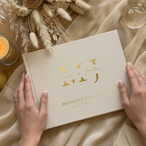 以下が含まれることがあります： クリーム色の結婚式のゲストブック、金色の箔文字。表紙には「Madison & Jonathan」と「Brooklyn Botanic, NY 25 September 2025」と書かれています。本は開かれており、両手で持たれています。背景にはキャンドルと花があります。