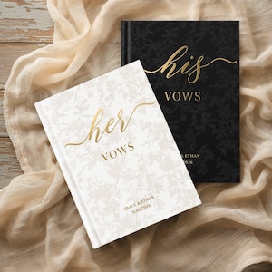 Op de afbeelding: Twee trouwgeloftenboeken. Eén is wit met een bloemmotief en gouden tekst met de tekst "her vows". De andere is zwart met gouden tekst met de tekst "his vows". De boeken liggen op een crèmekleurige stof.