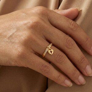 May include: A gold-colored ring with a heart-shaped padlock charm. The ring band is adorned with small, clear stones. The padlock charm has a keyhole design. The ring is on a person's finger.