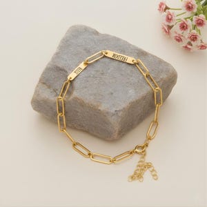 Könnte beinhalten: Goldfarbenes Kettenarmband mit rechteckigen Gliedern. Das Armband hat zwei rechteckige Stäbe mit den eingravierten Wörtern "RESIST" und "FREEDOM". Das Armband wird auf einem grauen Stein präsentiert.