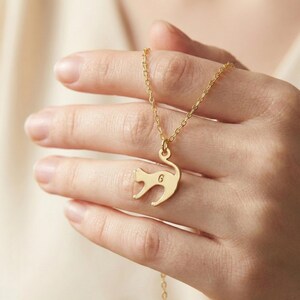 May include: A gold-colored necklace featuring a cat pendant with the letter "G" engraved on it. The pendant is suspended from a delicate chain. The necklace is displayed against a soft, neutral background.