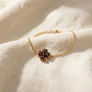 May include: A gold-colored bracelet featuring a four-leaf clover charm. The clover is made of four dark red gemstones set in a gold-colored frame. The bracelet is resting on a cream-colored fabric.