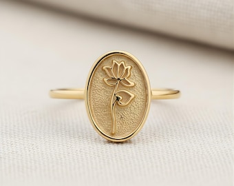May include: A gold-colored ring featuring an oval-shaped face with a detailed lotus flower design. The ring's band is thin and smooth, and the lotus flower is depicted in a delicate, engraved style. The ring is set against a soft, off-white background.