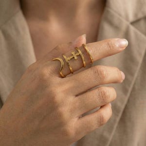 May include: A set of gold-colored rings, including a cross ring, worn on a finger. The rings have a minimalist design, with a smooth, polished finish. The rings are stacked together, creating a layered effect. The background is a neutral color.