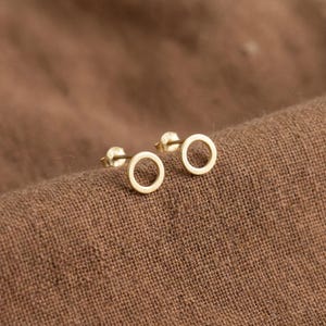 May include: A pair of small, gold-colored circle stud earrings. The earrings are displayed on a brown, textured fabric. The earrings have a simple, minimalist design, with a post and backing.