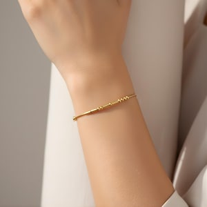 Könnte beinhalten: Ein zartes goldenes Armband mit einer Reihe kleiner, gleichmäßig verteilter Perlen und rechteckigen Stäben. Das dünne, elegante Armband hat einen einfachen Verschluss. Der Schmuck wird am Handgelenk getragen und zeigt sein minimalistisches Design.