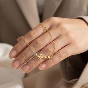 Könnte beinhalten: Eine zarte goldene Halskette mit dem Namen "Sarah" in Schreibschrift. Ein kleiner blauer Edelstein akzentuiert den Namen. Die Halskette wird präsentiert, um ihr Design und ihre Handwerkskunst hervorzuheben.