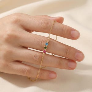 Könnte beinhalten: Ein zartes goldenes Armband mit einer feinen Kette, das eine Reihe farbiger Edelsteine ​​aufweist. Das Armband ist mit rosa, grünen, blauen und pfirsichfarbenen Steinen verziert. Das Armband wird auf einer Hand präsentiert.