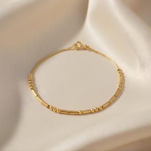 May include: A delicate gold bracelet featuring a fine chain and a series of small, cylindrical and spherical beads. The bracelet is displayed on a soft, off-white fabric, creating a minimalist and elegant aesthetic.