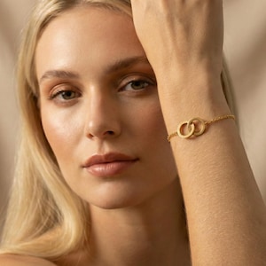 May include: Gold-toned bracelet featuring two interlocking circles. The delicate chain bracelet is displayed on a wrist, showcasing the jewelry's design. The bracelet's simple yet elegant design makes it a versatile accessory.