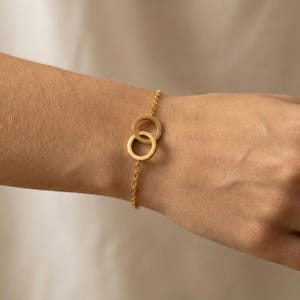 Könnte beinhalten: Ein goldfarbenes Armband mit zwei ineinandergreifenden Kreisen als zentrales Designelement. Das Armband hat eine feine Kette und wird an einem Handgelenk gezeigt. Die Kreise sind glatt und haben ein mattes Finish.