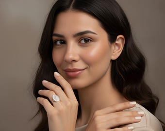May include: A silver ring with an oval-shaped face featuring a floral design. The ring is worn on a finger. The background is a solid, neutral color.