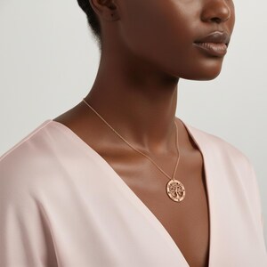 May include: A rose gold necklace featuring a circular pendant with a tree of life design. The pendant hangs from a delicate chain. The necklace is worn against a light pink satin fabric.