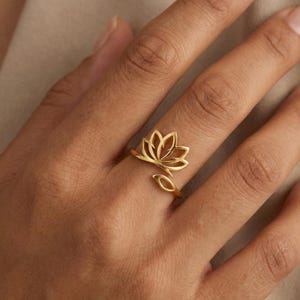 May include: A gold-colored ring featuring a lotus flower design. The ring has an open design with the lotus flower at the top and a leaf-shaped accent on the side. The ring is worn on a finger.
