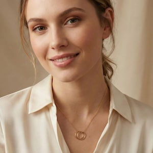 Peut inclure: Une femme porte un collier en or avec deux cercles entrelacés. Le collier est délicat et se porte sur une blouse crème. La femme sourit, et l'arrière-plan est beige neutre.