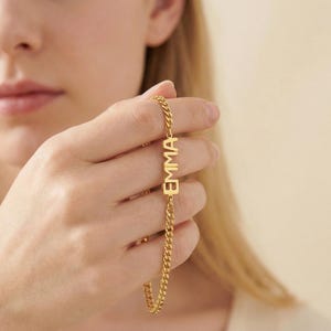 Könnte beinhalten: Ein goldfarbenes Kettenarmband mit dem Namen "EMMA" in einzelnen Buchstaben. Das Armband hat ein Panzerketten-Design und wird vor einem unscharfen Hintergrund gehalten.