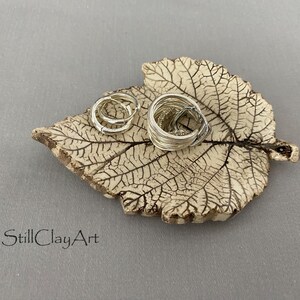 Può includere: Un piattino in ceramica color crema a forma di foglia con venature marrone scuro, contenente due anelli color argento. Il testo StillClayArt è stampato in nero nell'angolo in basso a sinistra. Il piattino presenta una superficie ruvida e irregolare.