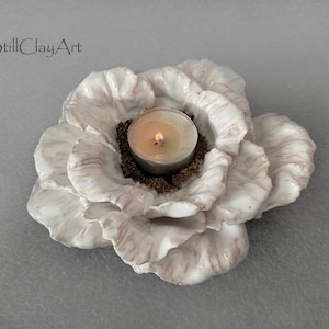 Puede incluir: Un portavelas de cerámica blanca con forma de flor, con pétalos superpuestos y ondulados que presentan vetas marrones sutiles. Una pequeña vela de té encendida se sitúa en el centro, rodeada de material granular marrón. El texto StillClayArt es visible.