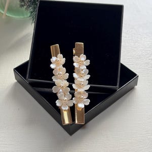 May include: Two gold-coloured barrettes, each decorated with white flower and pearl accents, are presented in an open black box. The rectangular barrettes feature a floral design, ideal for adding a touch of elegance to any hairstyle.