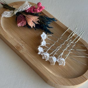 Könnte beinhalten: Ein Set silberfarbener Haarnadeln mit kleinen weißen Rosenverzierungen an den Enden. Die Nadeln sind in einer Gruppe angeordnet und zeigen die metallische Drahtstruktur sowie das detaillierte Blumendesign jeder einzelnen weißen Rose.