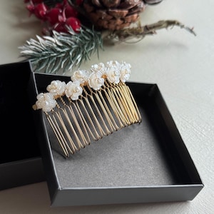 Puede incluir: Un peine para el cabello dorado con detalles de flores blancas, presentado en una caja negra. El peine tiene múltiples púas y está adornado con pequeños y detallados adornos florales. El diseño sugiere un accesorio nupcial o para ocasiones especiales.