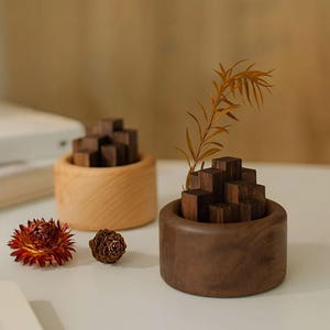 Op de afbeelding: Twee houten decoratieve vazen. Eén is lichtgekleurd, de andere is donkerbruin. Elke vaas heeft een cilindrische vorm met een cluster van kleine, donkere houten blokken erin. Een gedroogde bloem en een dennenappel staan op de voorgrond.