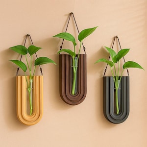 May include: Three wall-mounted planters with green plants. Each planter has a wooden, arched design in yellow, brown, and black. A clear glass tube holds the plant stems. The planters hang from a brown cord.