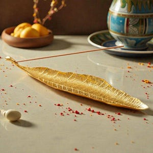 May include: A gold-colored leaf-shaped incense holder with detailed veining. An incense stick rests on the holder, with a small white object and scattered red and yellow granules nearby. A decorative bowl with fruit and a patterned vase are in the background.
