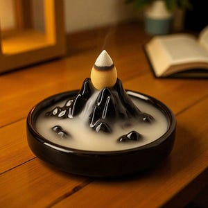 May include: A black ceramic incense burner shaped like a mountain range, with a cone-shaped incense stick releasing smoke. The burner sits on a wooden surface, creating a serene and decorative ambiance.