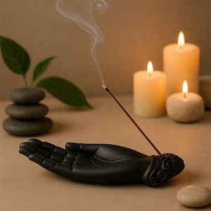 May include: A black ceramic hand-shaped incense holder with a burning incense stick. The holder is next to three lit candles and a stack of stones. The incense stick emits a thin stream of smoke.