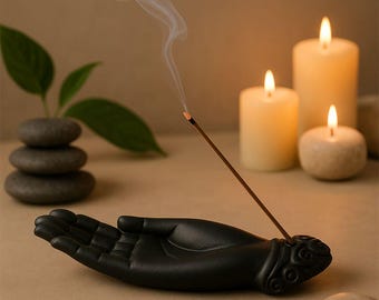 May include: A black ceramic hand-shaped incense holder with a burning incense stick. The holder is next to three lit candles and a stack of stones. The incense stick emits a thin stream of smoke.