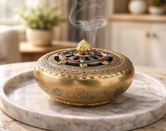 May include: A golden incense burner with intricate patterns and a decorative lid, emitting wisps of smoke. The burner is adorned with colorful beads and sits on a marble tray, creating a serene and decorative ambiance.
