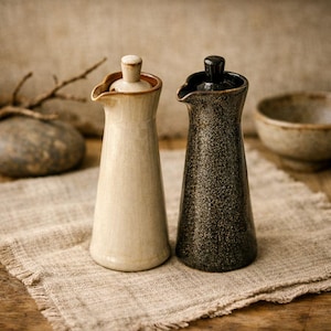 Op de afbeelding: Twee keramische olie-dispensers. Eén is crèmekleurig met een bruine rand en deksel, de andere is zwart met een gespikkelde textuur en een zwart deksel. Beide hebben een schenktuit en staan op een textiele stof met neutrale kleur.
