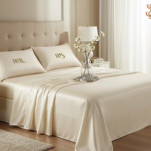 Könnte beinhalten: Ein cremefarbenes Bettwäsche-Set mit den Worten "HER" und "HIS" auf den Kissenbezügen. Das Bett ist mit einem glatten, seidigen Stoff bezogen. Eine Vase mit weißen Blumen steht auf einem silbernen Tablett auf dem Bett. Das Kopfteil ist beige.