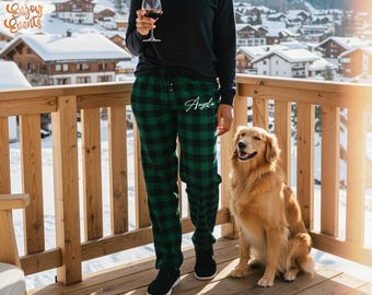 May include: Green and black plaid pajama pants with the name "Angela" in white script. A person wearing a black long-sleeve shirt and black shoes is holding a glass of red wine. A golden retriever dog sits on a wooden deck.