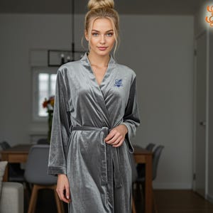 May include: A grey velvet dressing gown with long sleeves and a matching belt. The gown has a wrap-around design and a small embroidered emblem on the chest. The robe is a soft, luxurious garment.