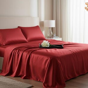 Könnte beinhalten: Ein Bett ist mit einem roten Satin-Bettwäsche-Set bezogen, einschließlich zweier Kissenbezüge. Ein schwarzes Tablett mit weißen Blumen liegt auf dem Bett. Eine weiße Lampe und eine kleine Uhr stehen auf dem Nachttisch.