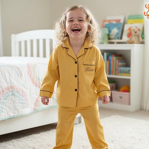 Può includere: Un bambino indossa un pigiama giallo senape con rifiniture nere e il nome "Rio" ricamato sulla camicia. Il pigiama ha una parte superiore abbottonata e pantaloni abbinati. Il bambino sorride in una camera da letto.