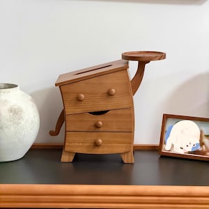May include: A quirky wooden tissue box with a distinctive design. The box features three drawers, a tray, and two curved wooden arms. The wood is a light brown colour. The tissue box is on a black surface.