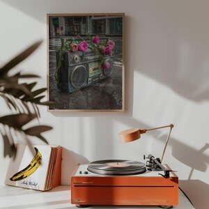 May include: A framed print featuring a vintage boombox adorned with pink flowers, set against a city street backdrop. A record player with a vinyl record and a lamp are in the foreground, with a book cover featuring a yellow banana.
