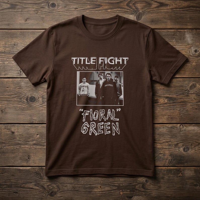 Puede incluir: Una camiseta de manga corta de color marr&oacute;n oscuro con un estampado gr&aacute;fico blanco. El dise&ntilde;o incluye el texto "TITLE FIGHT" sobre una fotograf&iacute;a rectangular en blanco y negro de cuatro personas, con "06 'FIORAL GREEN' 99" debajo.
